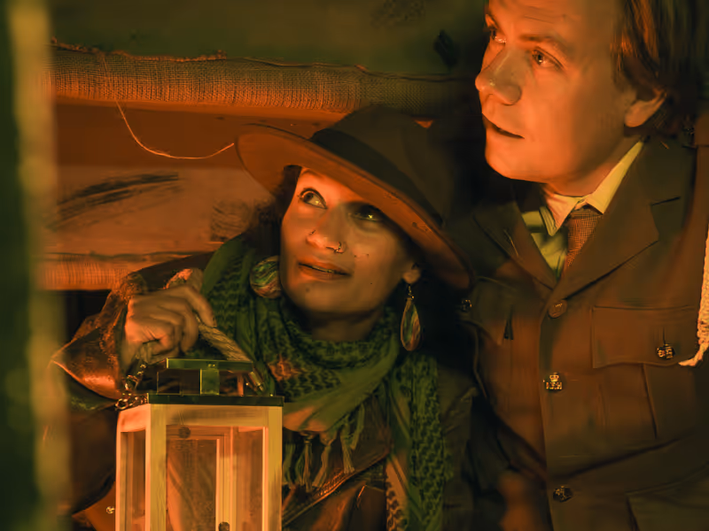 A woman in a hat and scarf holding a lantern looks up beside a man in a suit jacket in warm lighting.