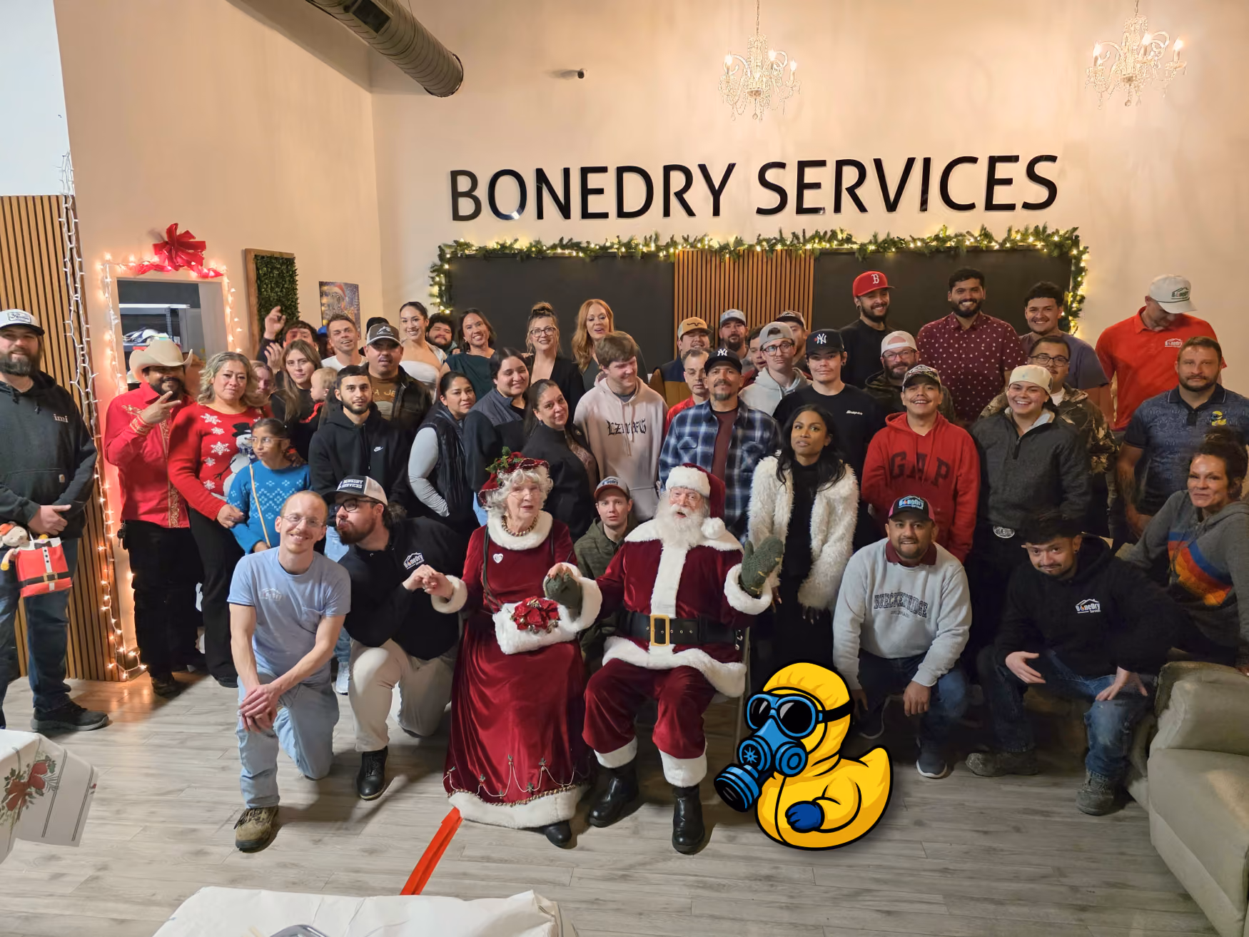 Large group of people posing indoors with Santa, Mrs. Claus, and holiday decorations at Bonedry Services.
