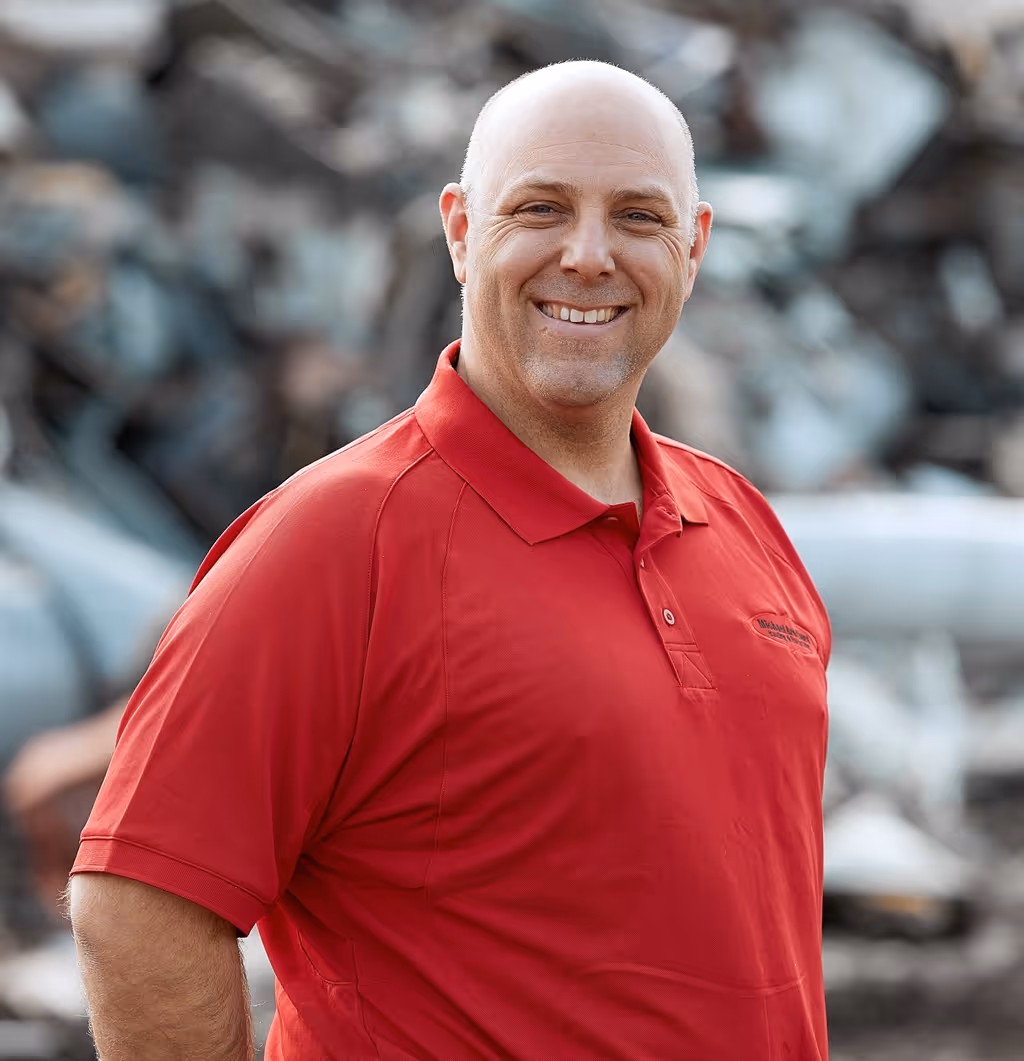 Tom wearing a red polo shirt standing outdoors with a blurred industrial background.