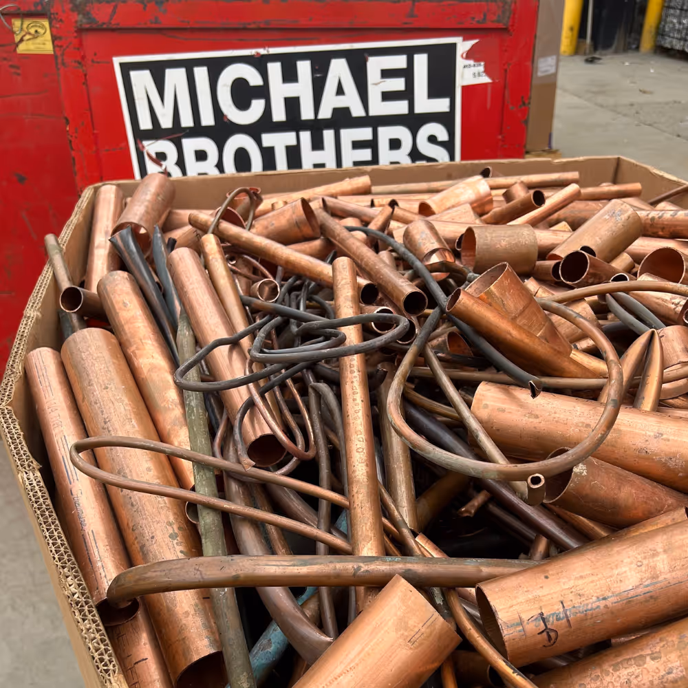 Cardboard container filled with various lengths and shapes of copper pipes and tubes with a red metal container labeled Michael Brothers in the background.