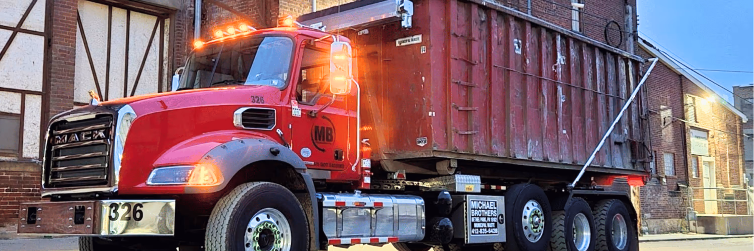 Red Mack dump truck with illuminated lights parked in front of brick buildings at dusk.