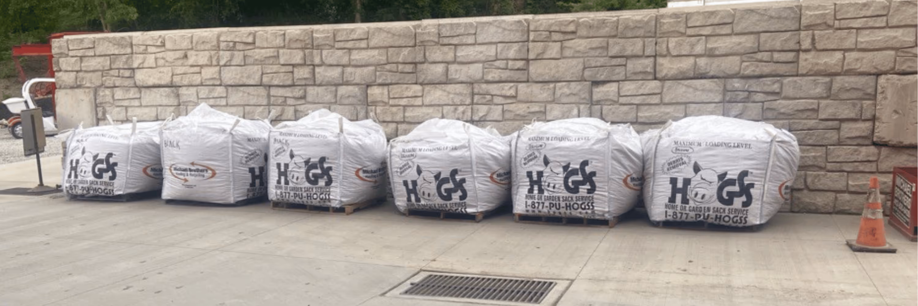 Six large white bags labeled with a pig logo and the text 'HOGS' stacked on pallets in front of a stone wall outdoors.