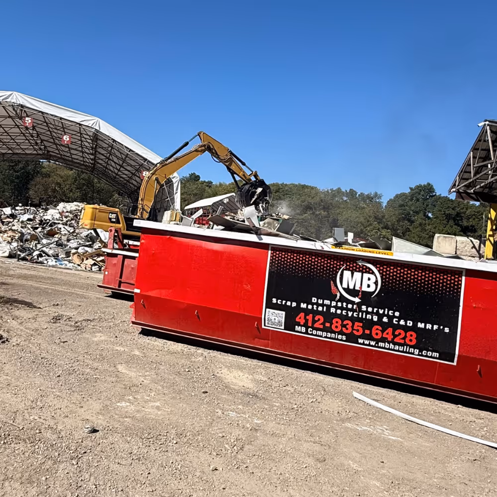 Yellow excavator loading debris into a large red dumpster labeled with MB Companies dumpster and scrap metal recycling service information.