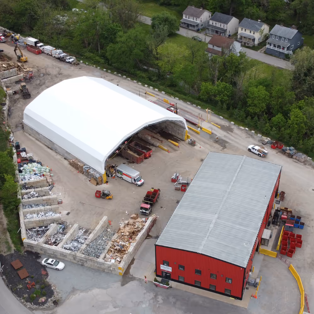 Aerial view of a recycling facility with a large white tent, red building, sorted material bins, trucks, and surrounding greenery.