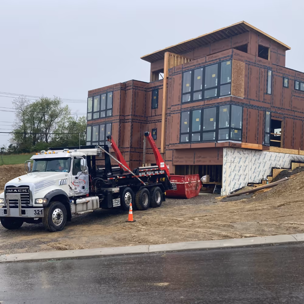 Construction site showing a modern house framed with wooden panels and large windows, alongside a white Mack truck parked on gravel with a red dumpster nearby.