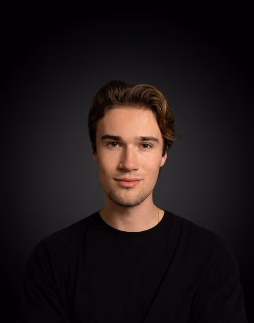 Porträt eines jungen Mannes mit braunem Haar, der ein schwarzes T-Shirt vor einem dunklen Hintergrund trägt.