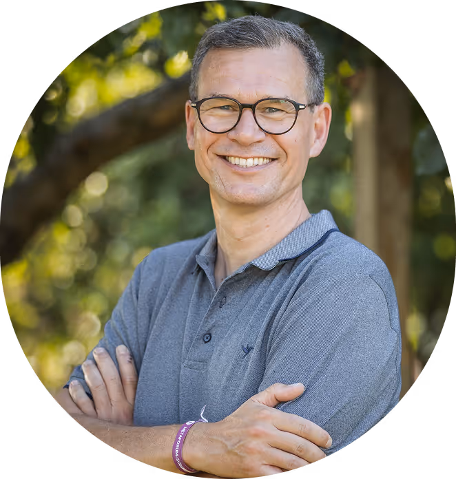 Mann mit grauem Polohemd und Brille lächelt mit verschränkten Armen vor unscharfem grünem Hintergrund.