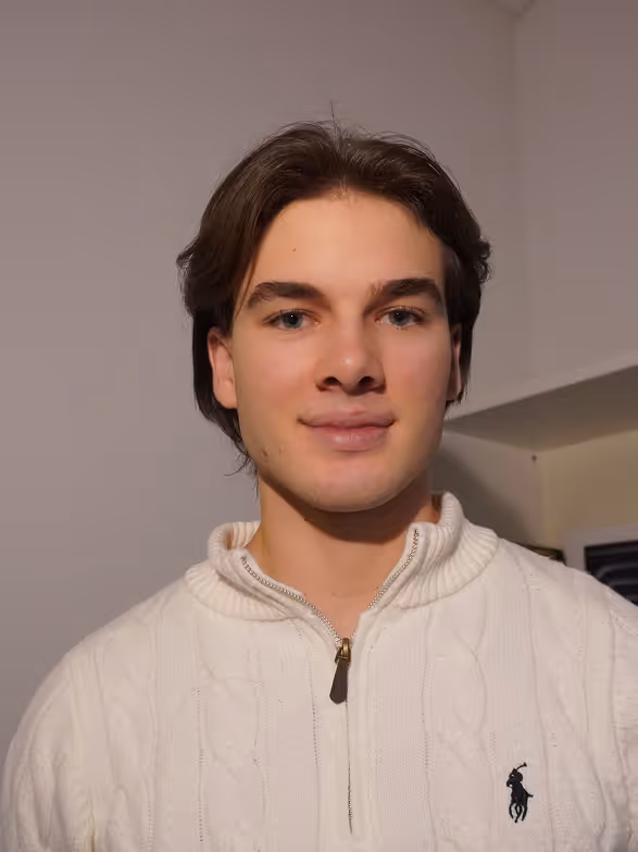 Junger Mann mit braunen Haaren in einem weissen Pullover mit Reissverschluss vor einem hellen Hintergrund.