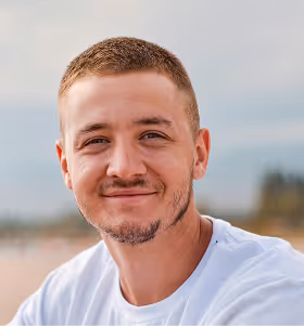 Junger Mann mit kurzem braunem Haar und leichtem Bart lächelt, trägt weißes T-Shirt, unscharfer Hintergrund mit Himmel.