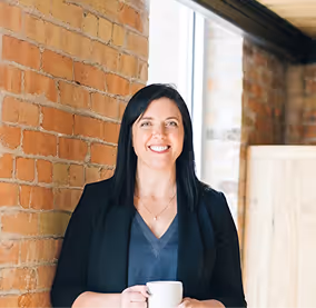 Frau mit schwarzen Haaren, die lächelnd eine Tasse hält und vor einer Ziegelsteinwand steht.