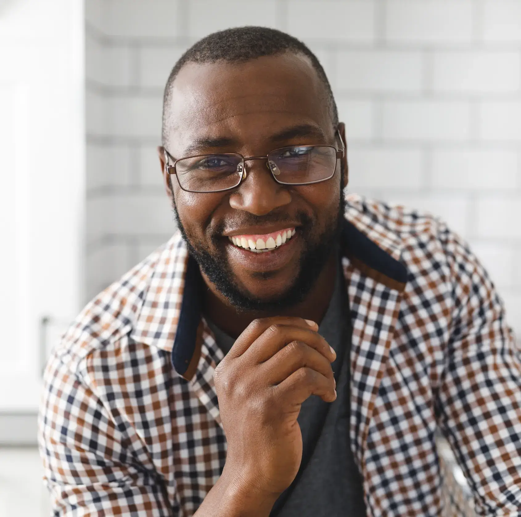 A man wearing glasses and a checkered shirt smiles while resting his chin on his hand.