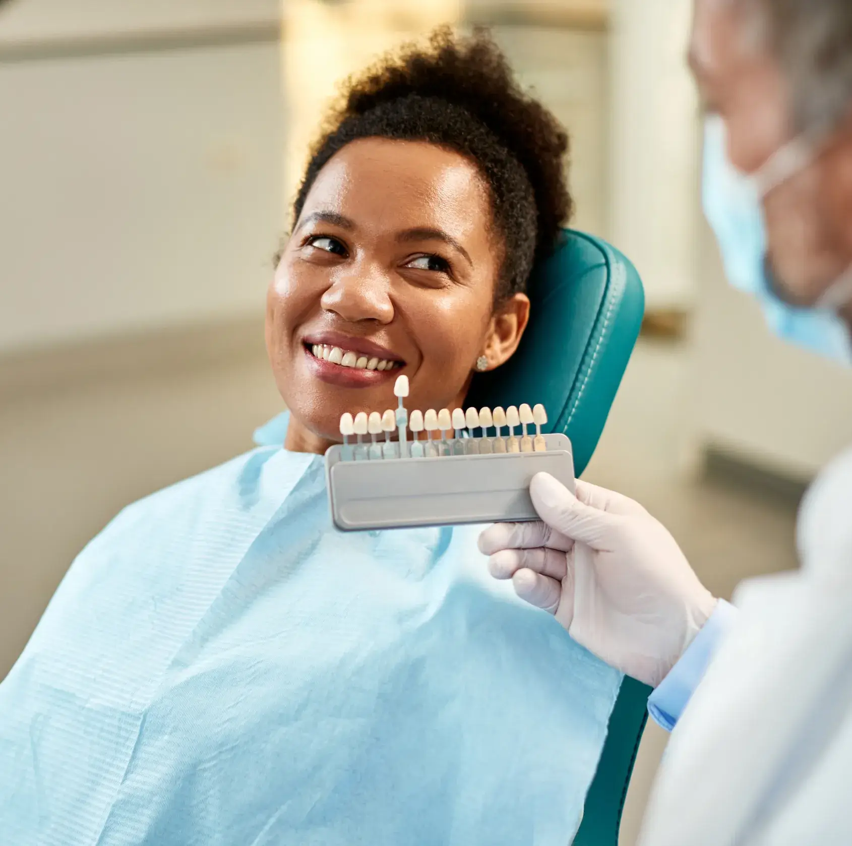 A woman is sitting in a dental chair, smiling as a dentist holds a shade guide.