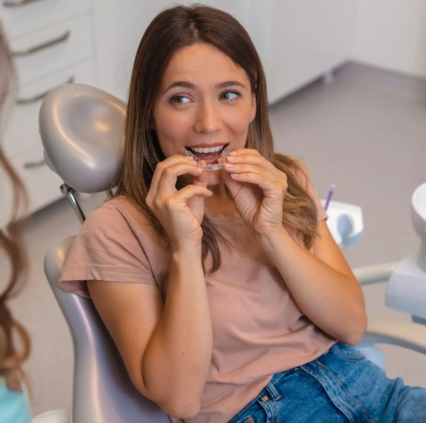 A woman sitting in a dental chair holds an Invisalign retainer near her mouth.