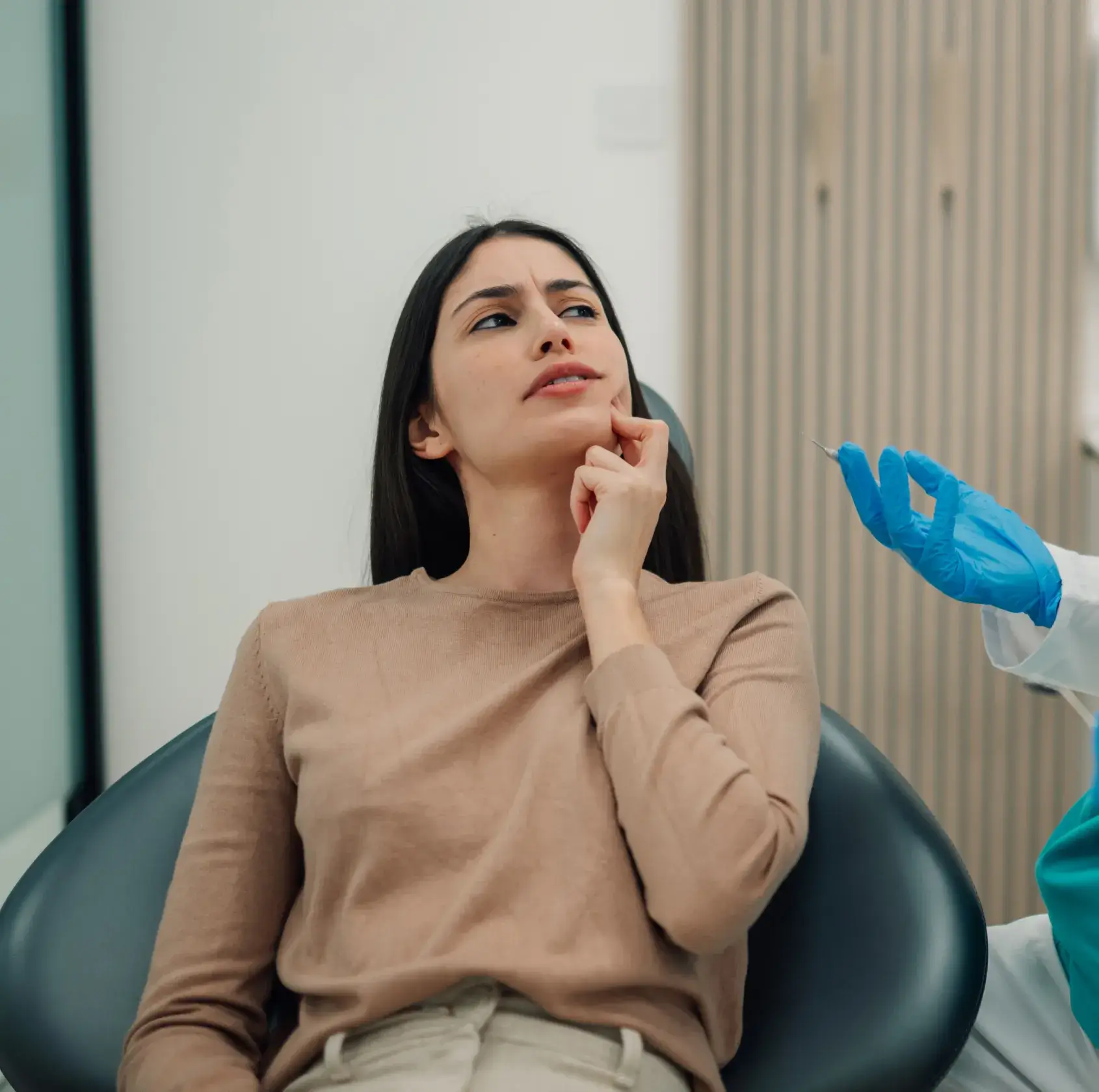 A woman sits in a dental chair, touching her cheek, while a dentist in gloves gestures.