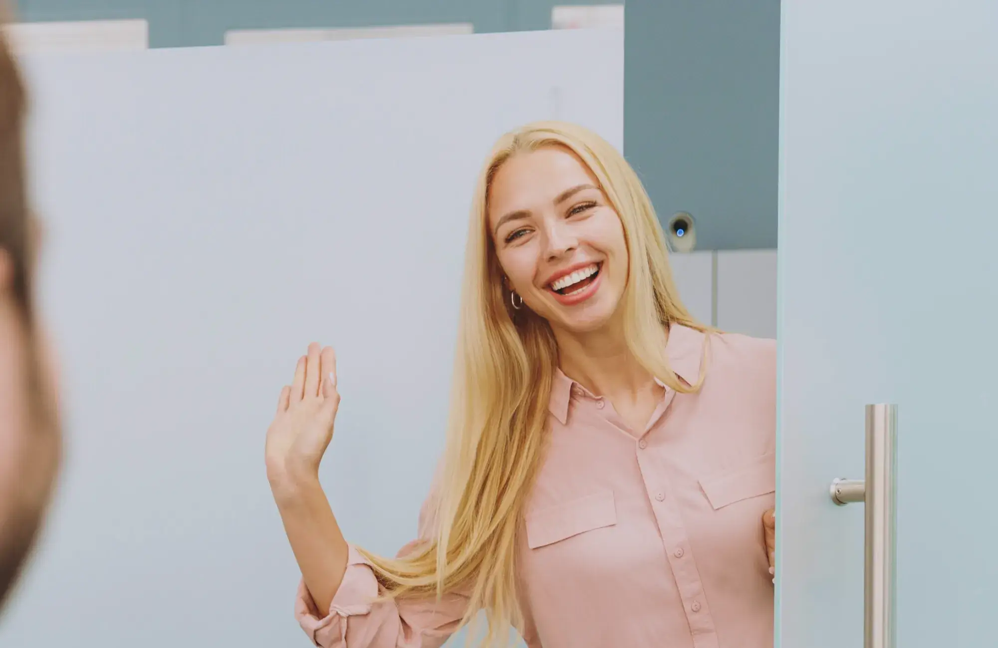 A woman in a pink shirt waves and smiles while standing in a doorway.