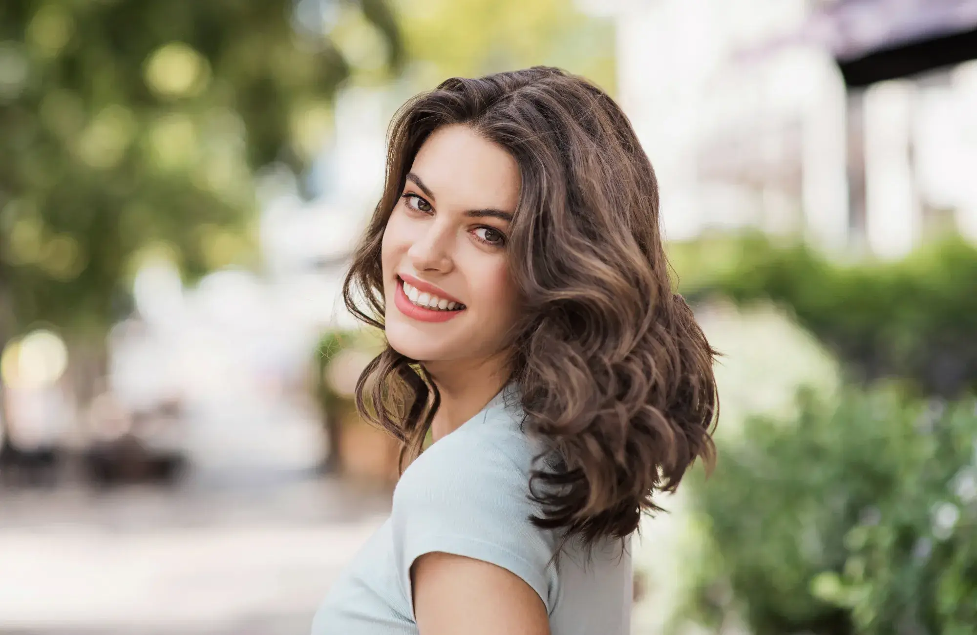 A woman with wavy brown hair smiles while standing on a sidewalk outside.