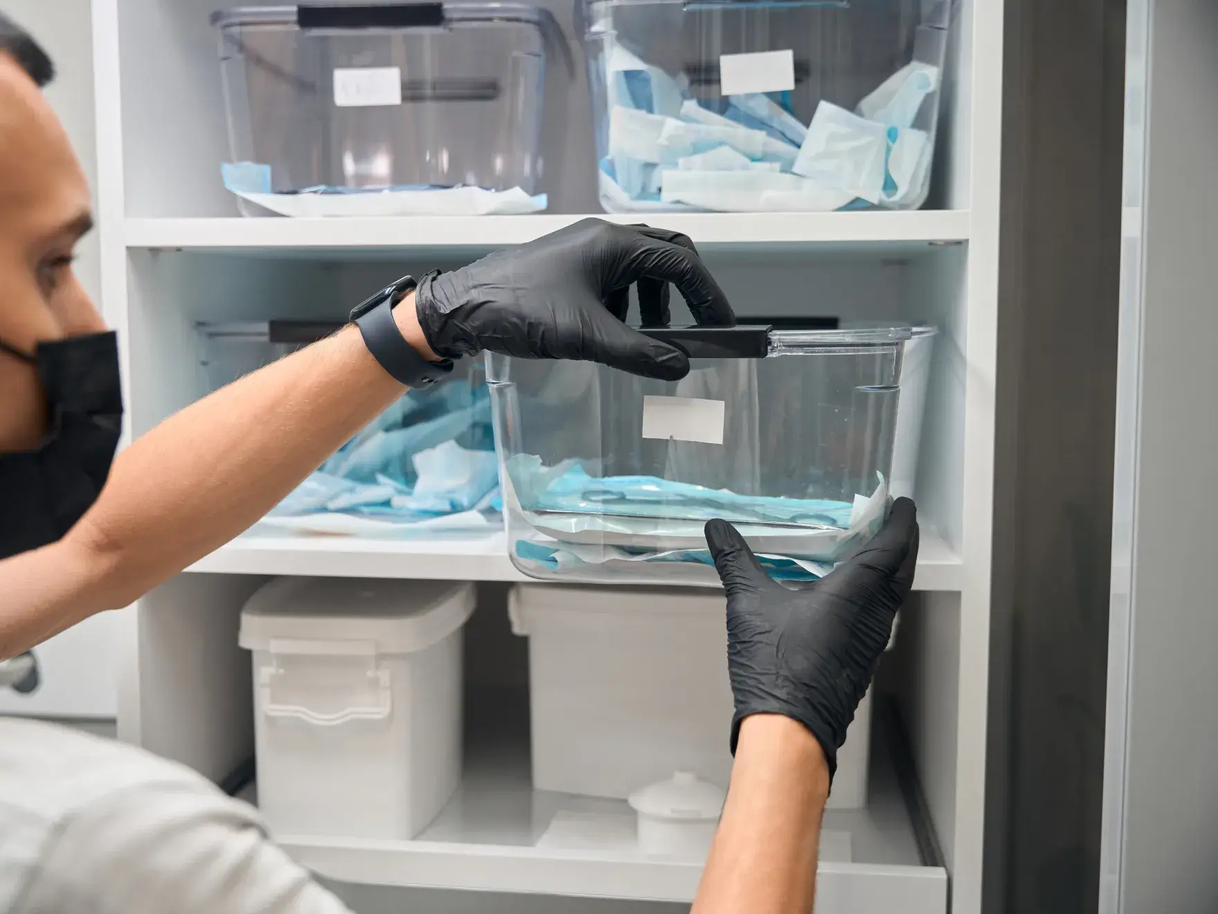 Person wearing gloves organizes plastic containers on shelves in a storage area.
