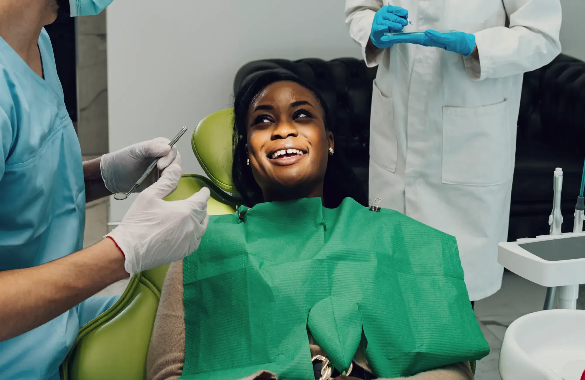 A woman smiles while sitting in a dental chair, surrounded by two dentists preparing tools.