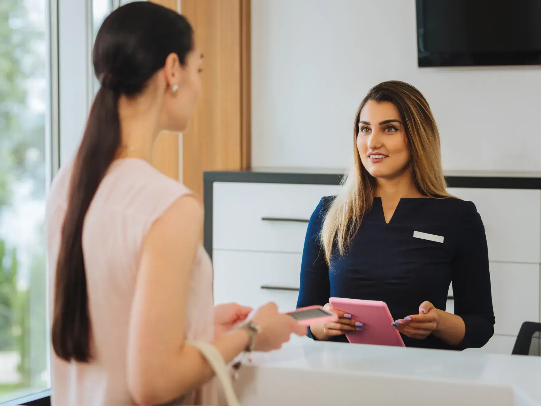 A woman at a reception desk assists a visitor holding a smartphone.