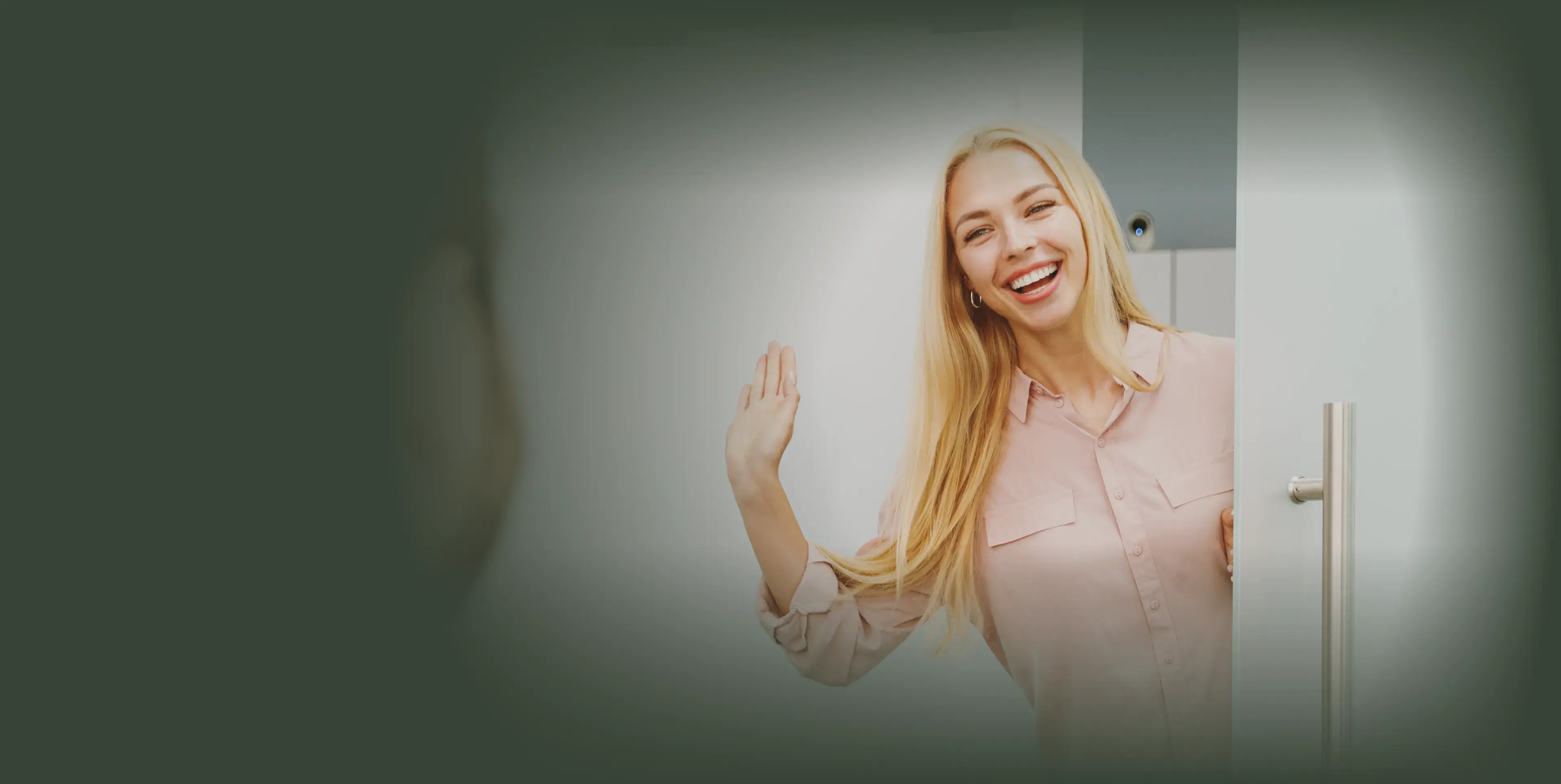 A woman with long blonde hair waves and smiles from a doorway.