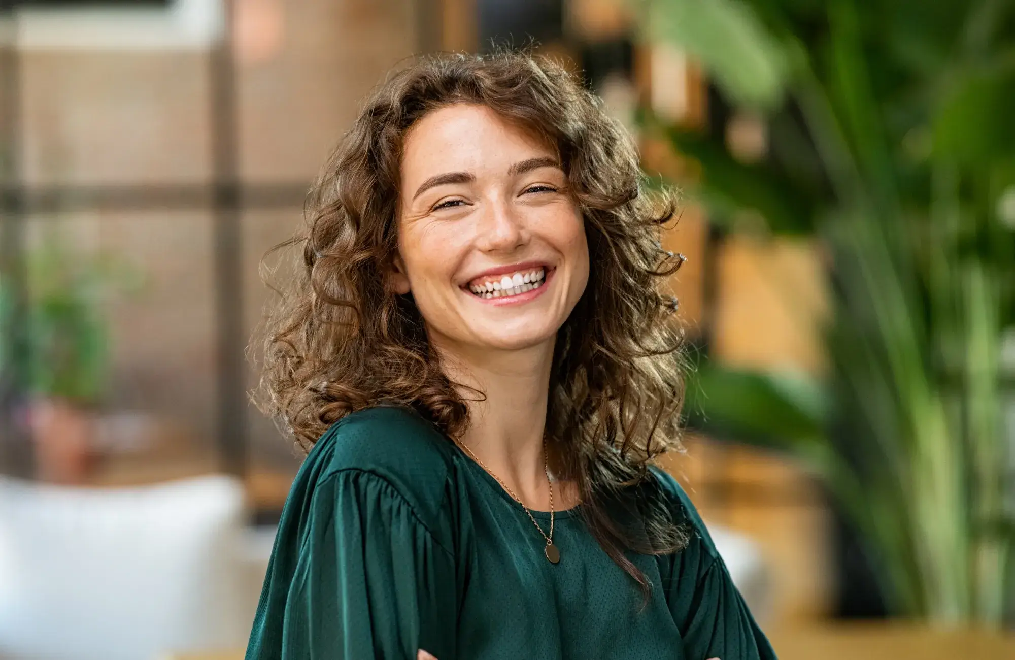 A person with curly hair smiles warmly while wearing a dark green shirt indoors.