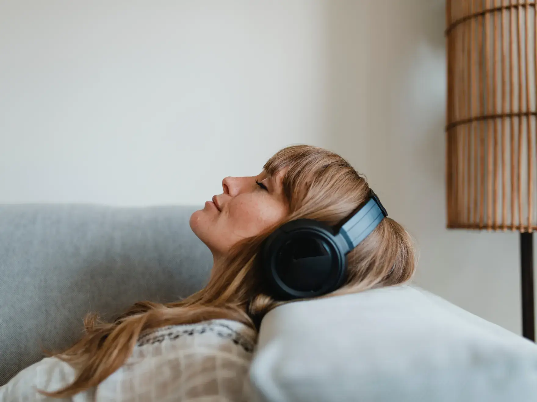 A woman wearing headphones leans back on a couch with eyes closed, appearing relaxed.