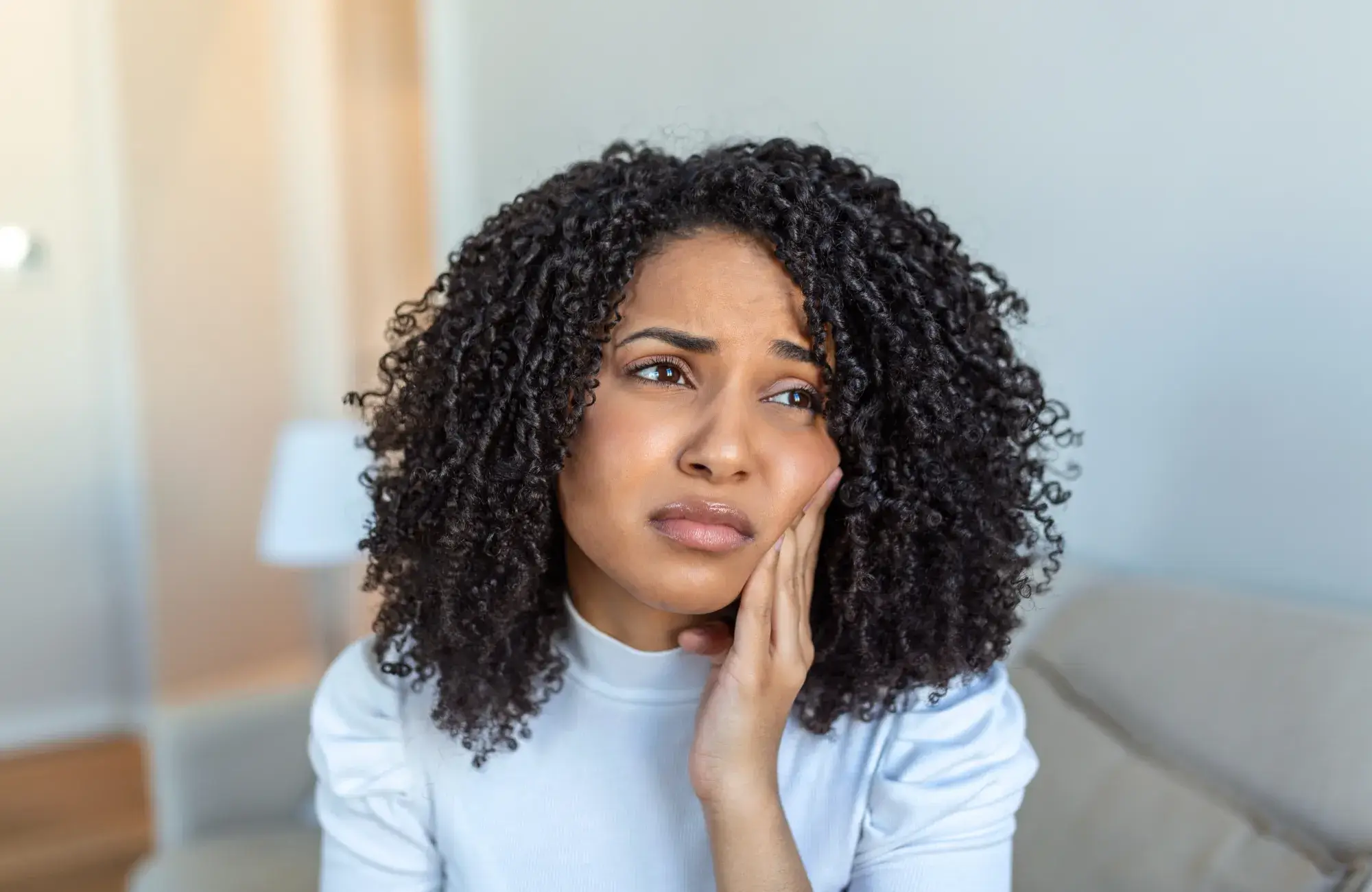 A woman with curly hair holds her cheek, appearing to feel discomfort or pain.