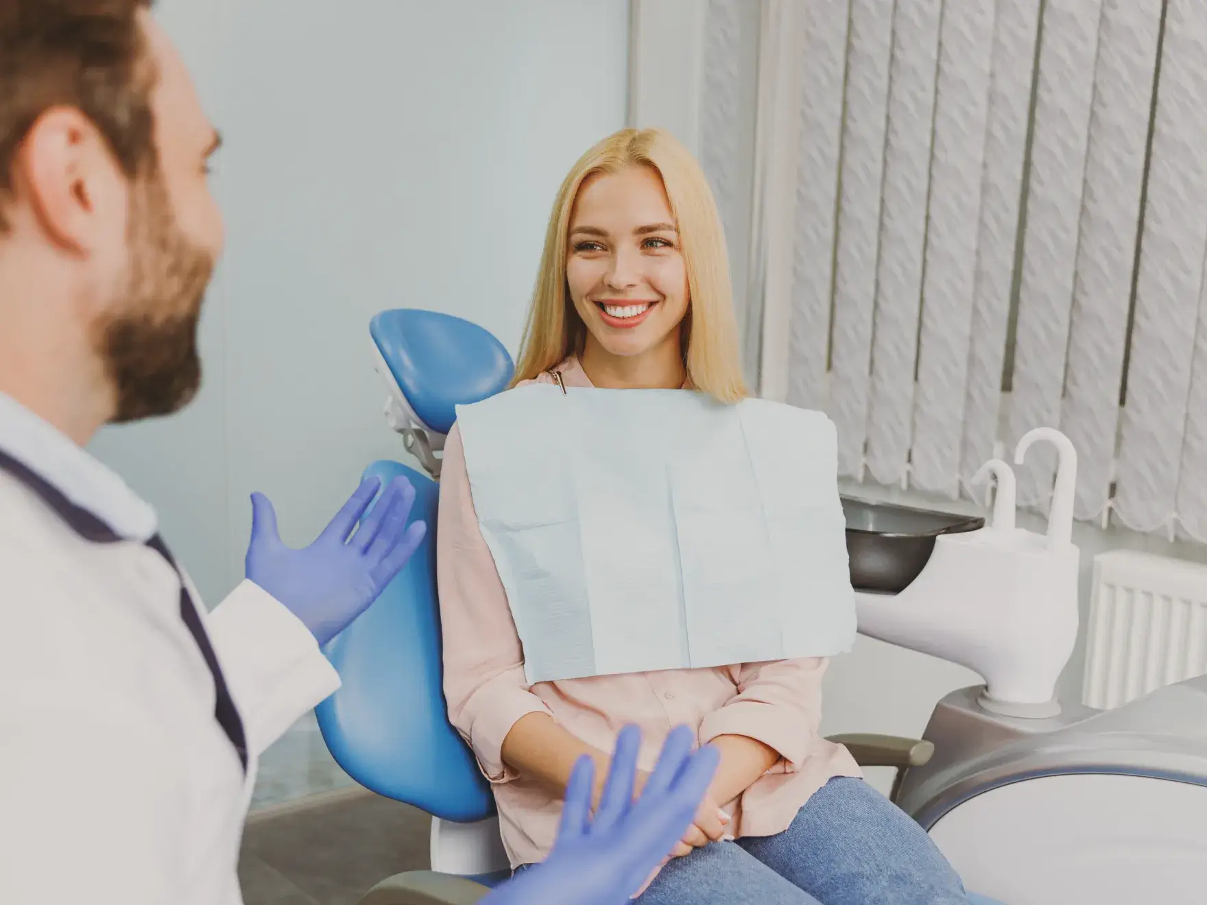 A dentist wearing blue gloves talks to a smiling woman sitting in a dental chair.