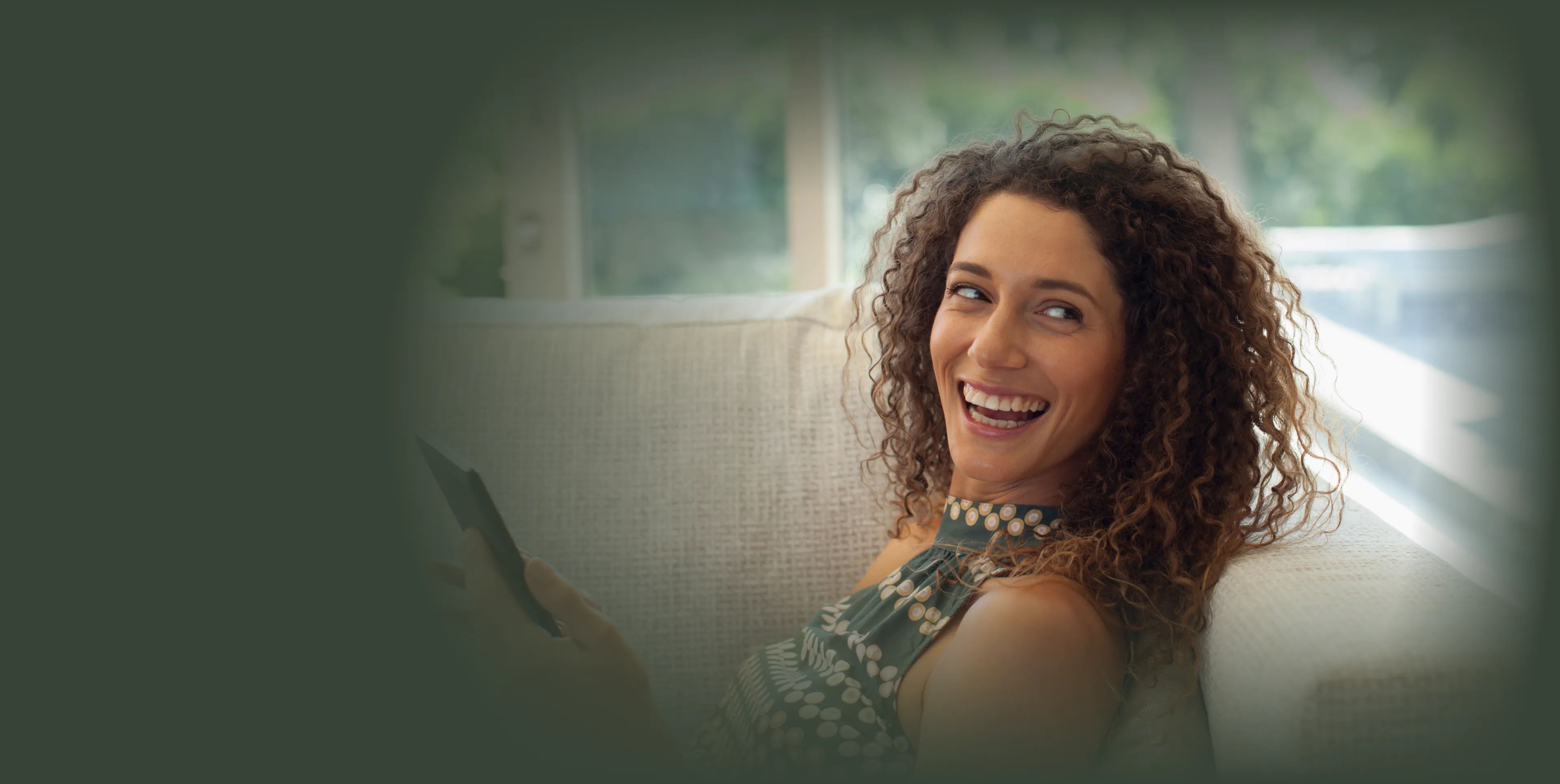 A person with curly hair sits on a couch, smiling and holding a smartphone.