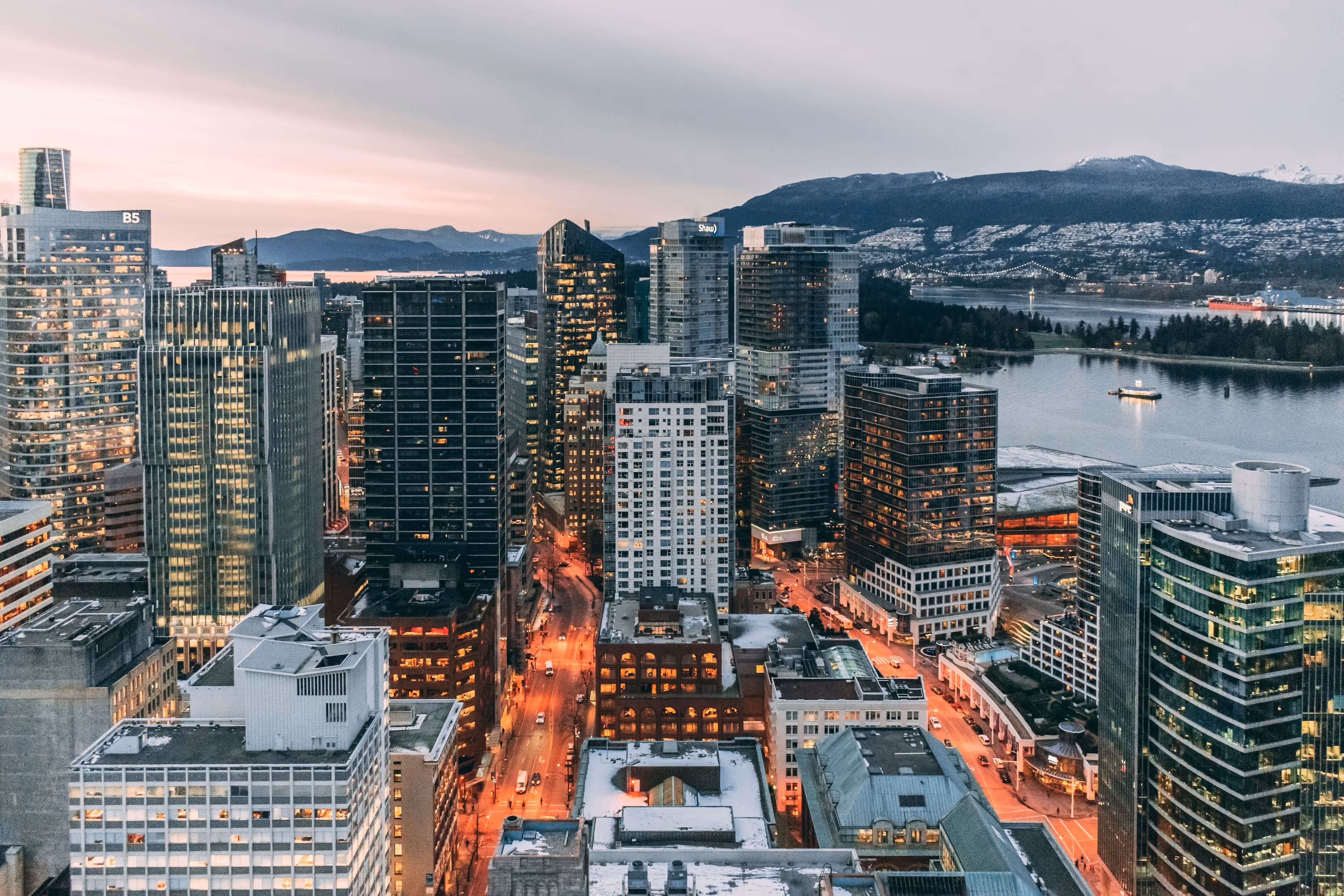 Evening cityscape with lit high-rises, snowy mountains, and boat on water.