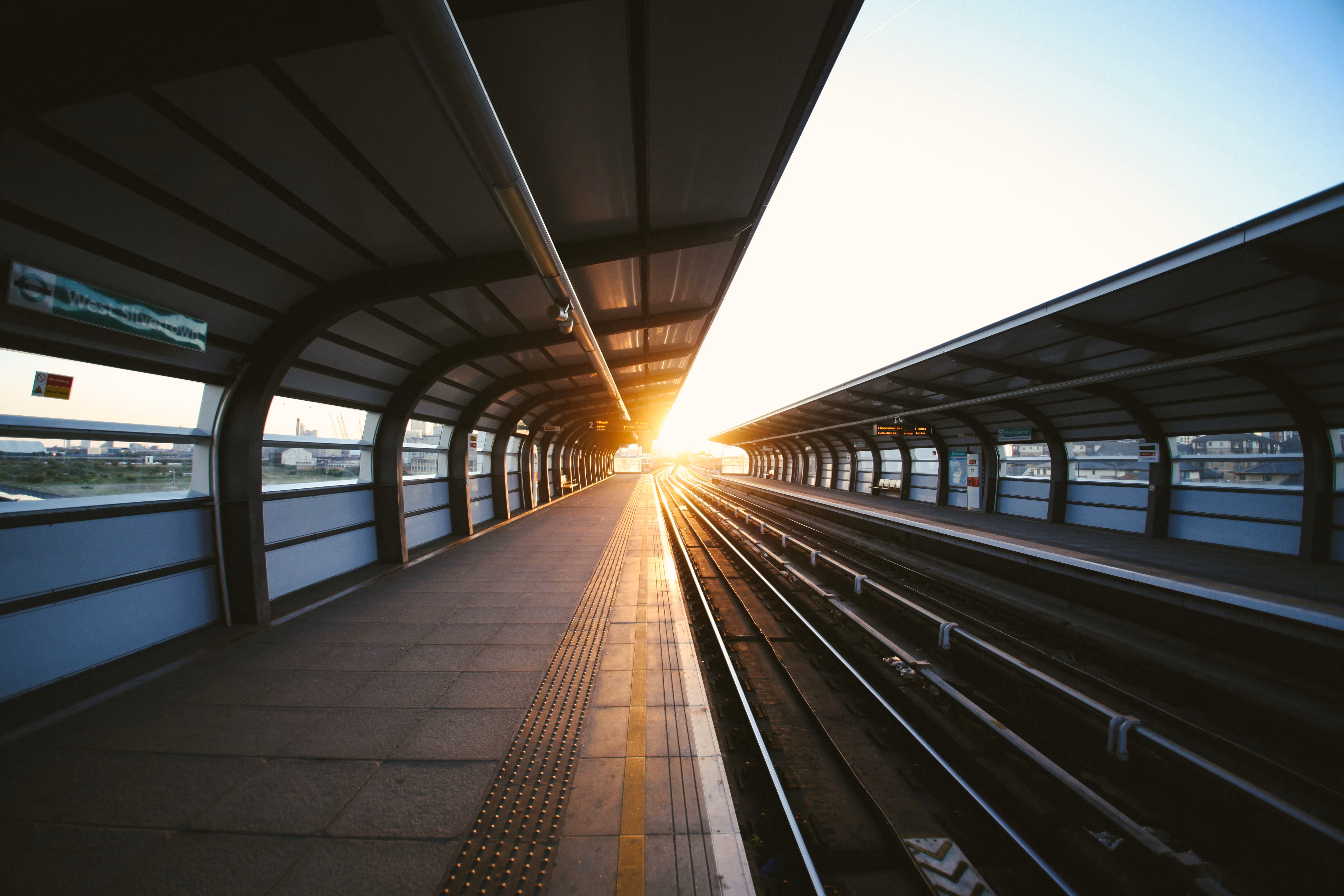 Modern train station platform with curved roof and warm sunrise or sunset lighting.