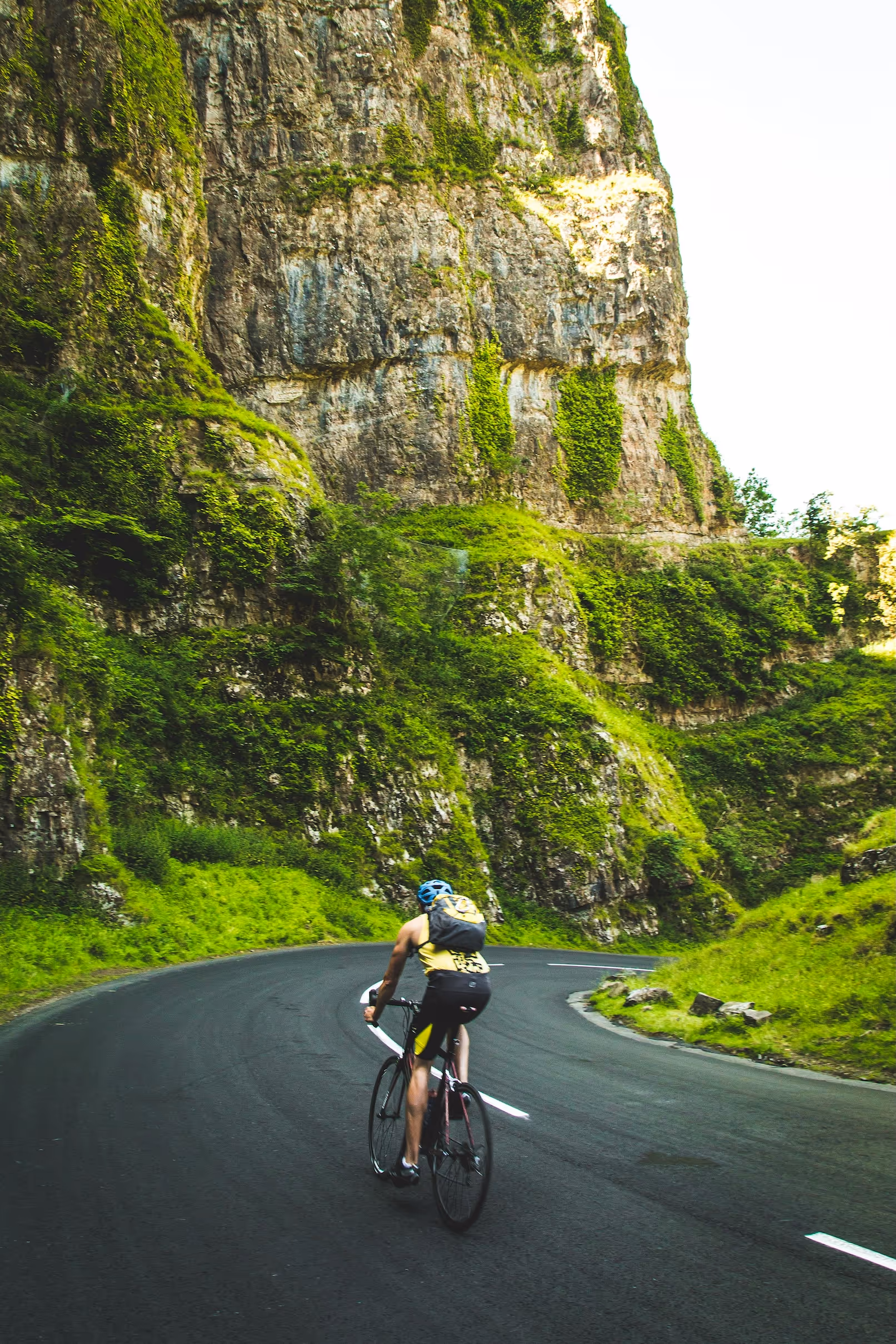 Cyclist on winding road through lush greenery and rocky cliffs.