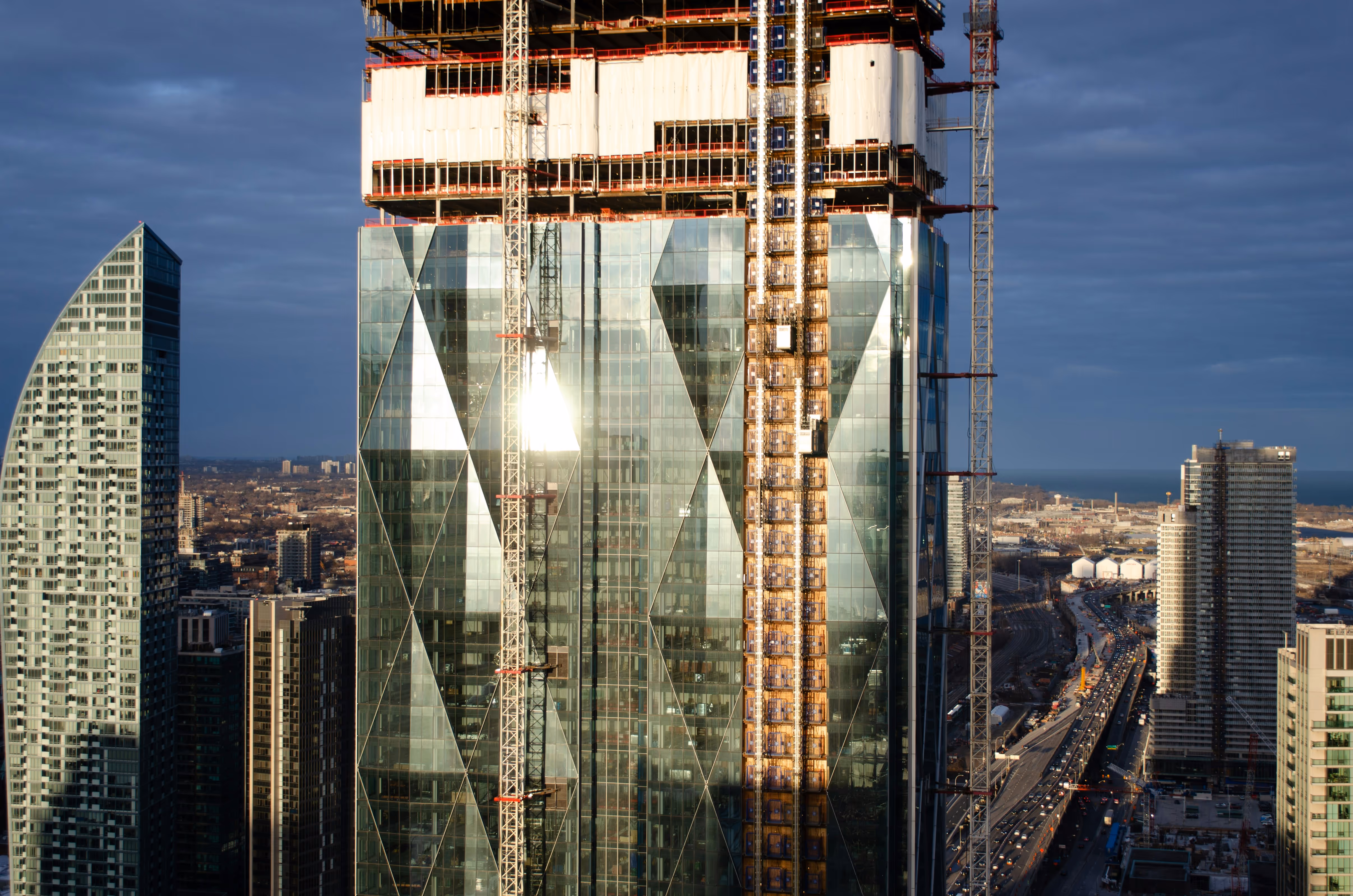High-rise building under construction with glass facade and cranes in cityscape.