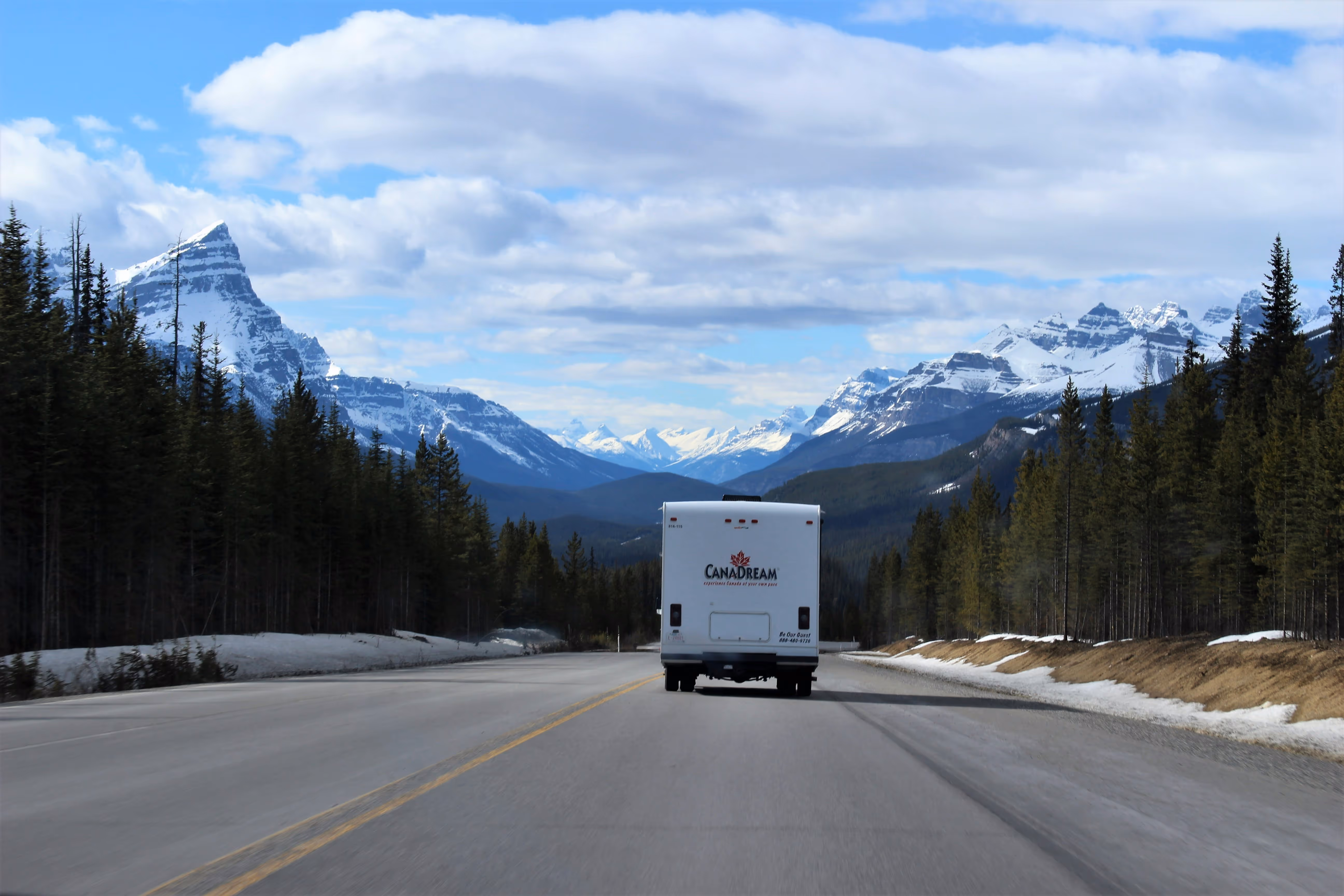RV on mountain road with snow-covered ground, trees, and peaks under cloudy sky.