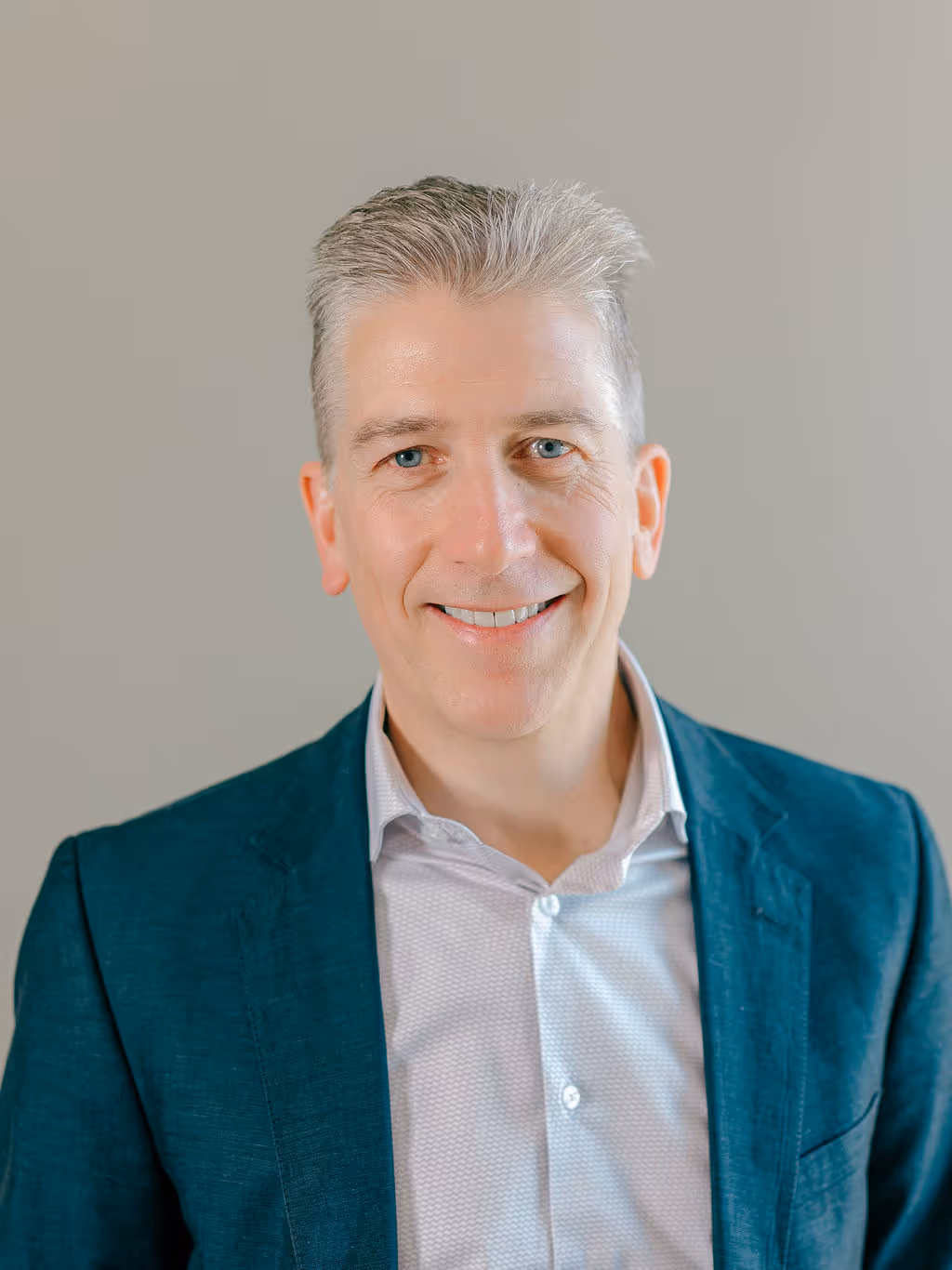 Smiling middle-aged man with gray hair wearing a dark teal blazer and white shirt against a plain gray background.