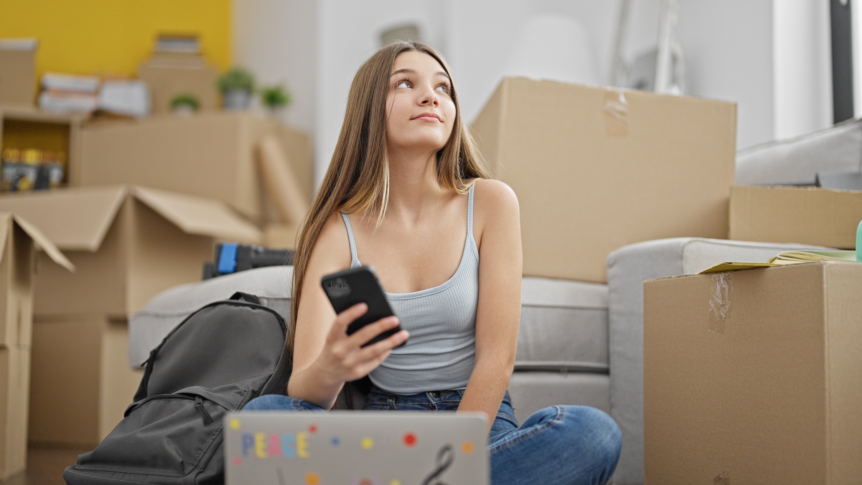 Student in apartment with moving boxes, computer, and a cell phone in hand