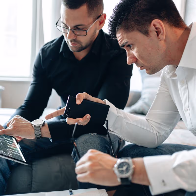 Two men seated on a couch, intently examining a tablet computer together.