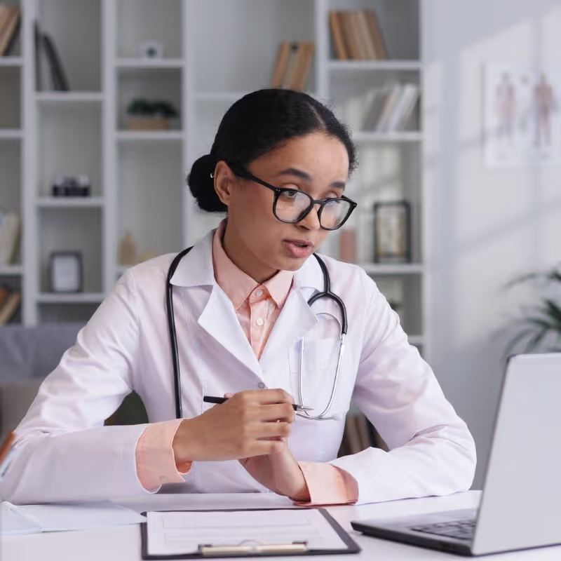 A woman in a white coat sits at a desk, focused on her laptop, surrounded by a professional workspace.