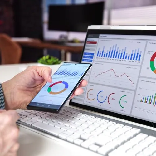 A person examines graphs on a laptop while holding a smartphone in their hand, showcasing multitasking with technology.