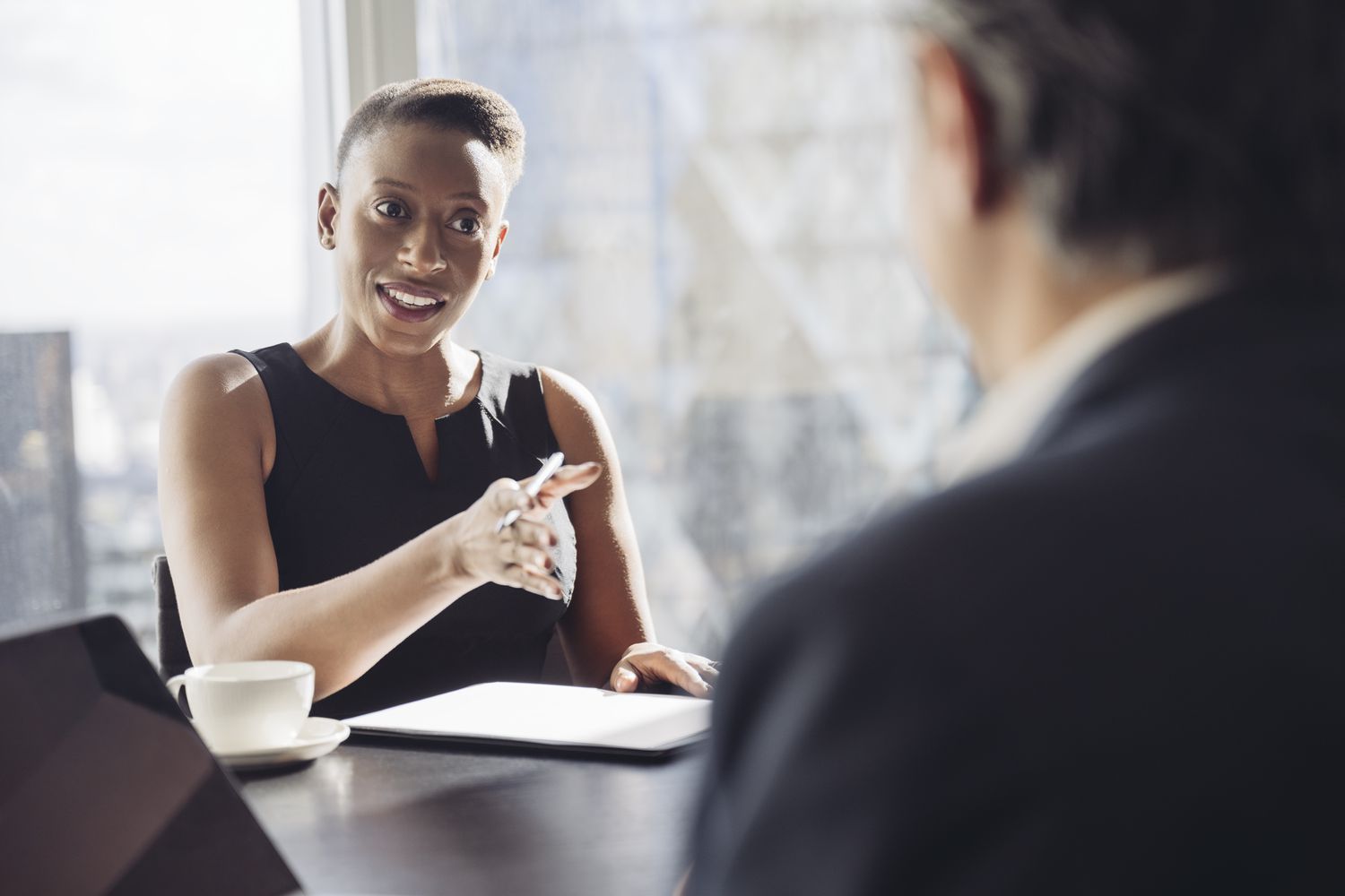 Woman in a sleeveless black dress gesturing while talking to a man in a suit across a table in an office with a city view.