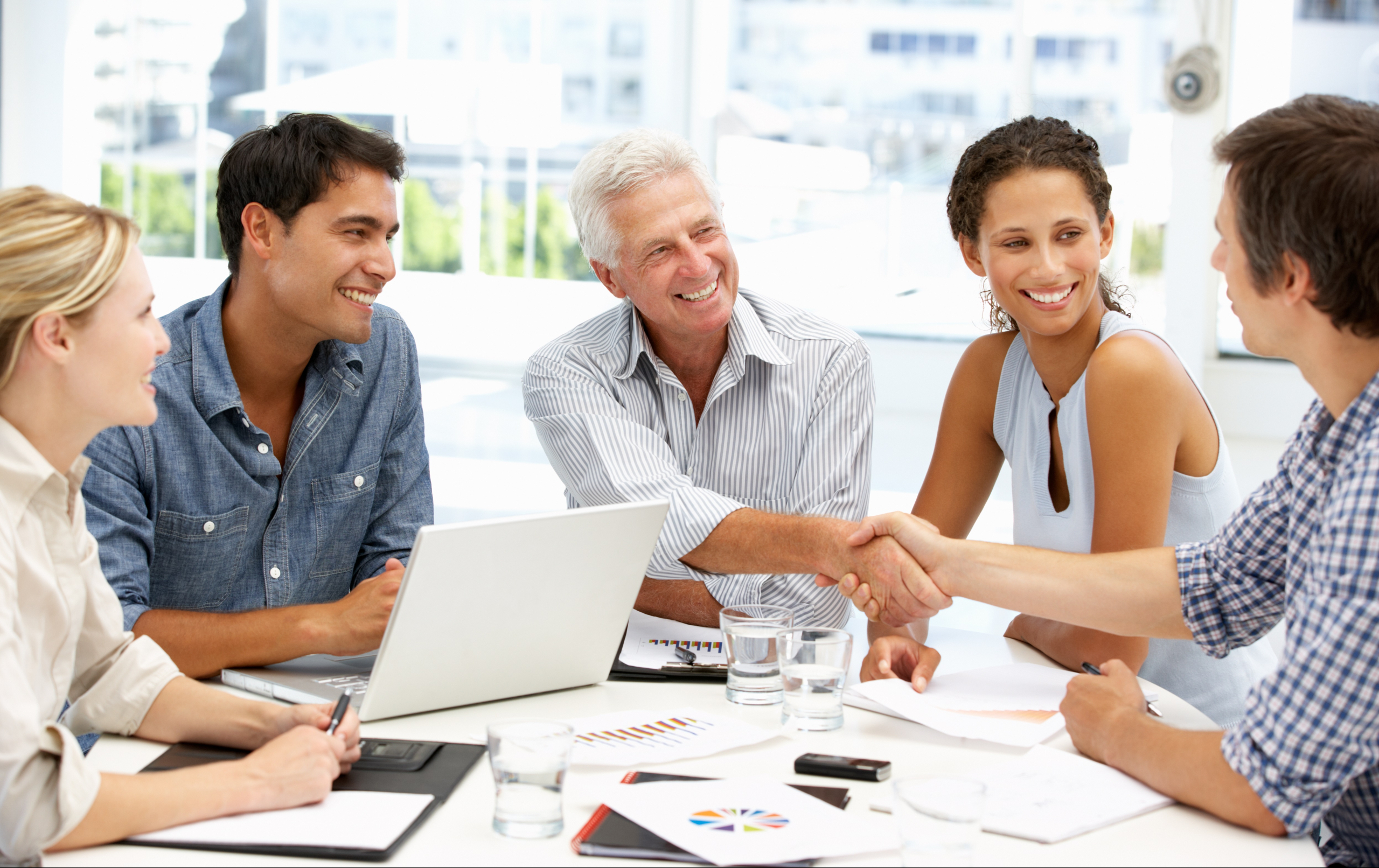 Five diverse colleagues smiling and shaking hands around a table with a laptop and documents in a bright office.
