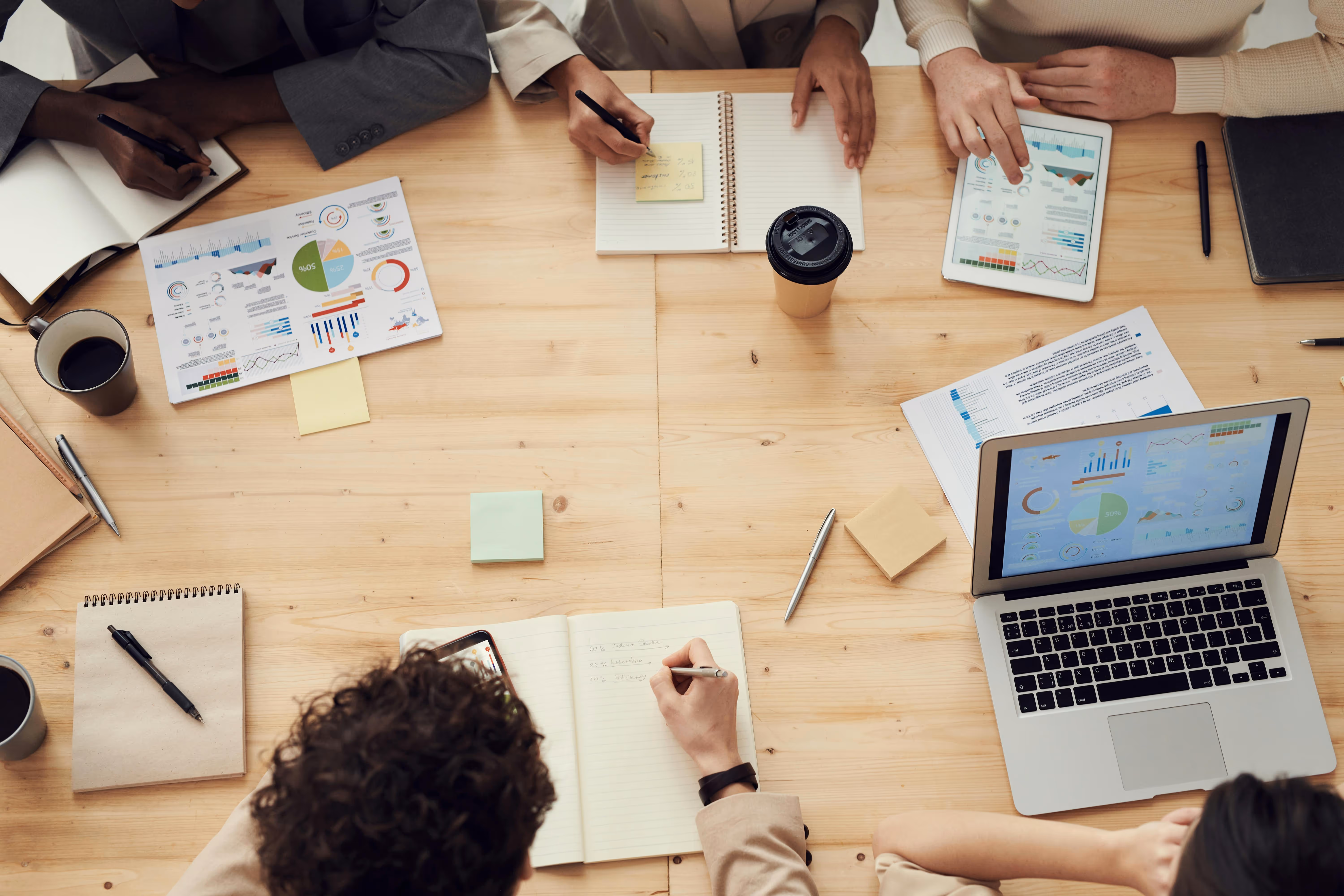 Overhead view of a group working at a wooden table with laptops, tablets, notebooks, charts, coffee cups, and sticky notes.