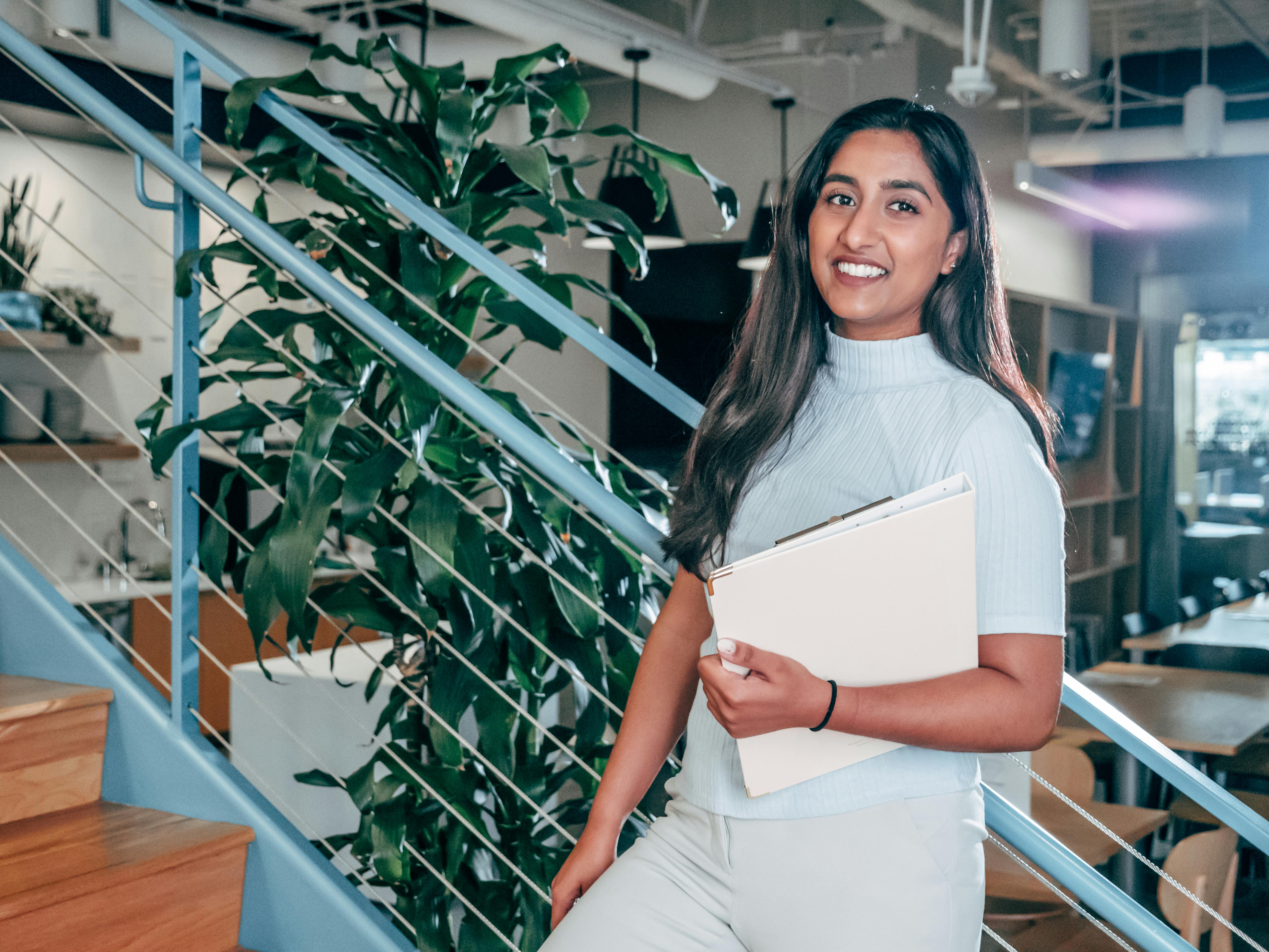 Smiling woman in light blue sweater holding a folder, standing by a staircase and indoor plant in a modern office.