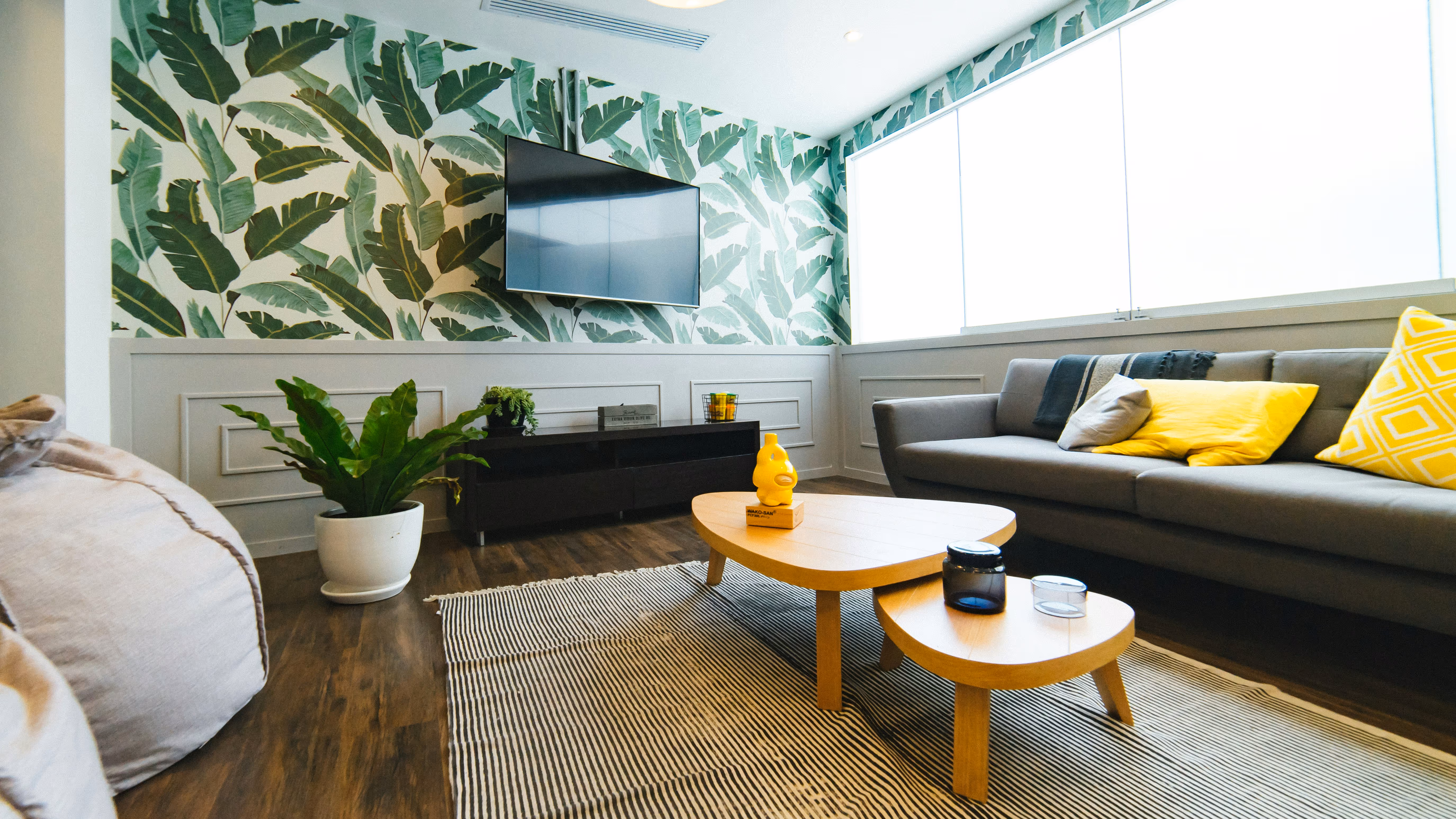 Modern living room with tropical leaf wallpaper, a mounted flat-screen TV, gray sofa with yellow pillows, wooden coffee tables, and a potted plant.