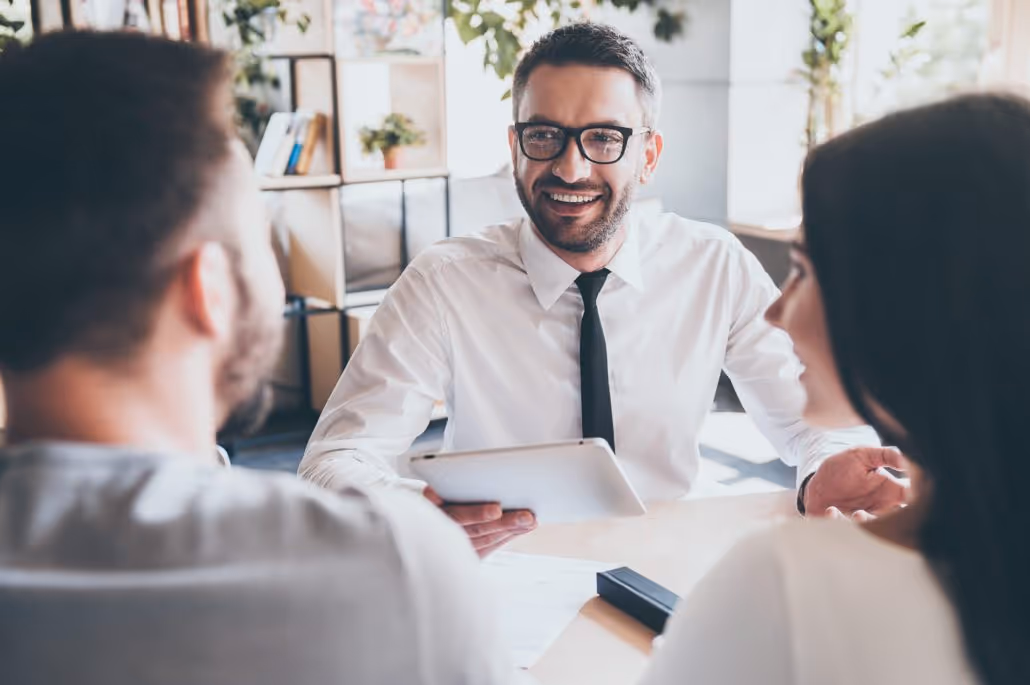 Smiling male advisor in glasses holding a tablet while talking to a couple at a desk in an office.