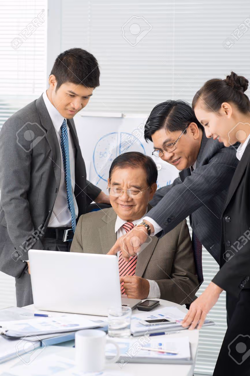 Four business professionals in suits gathered around a laptop, discussing work with charts and documents on the table.