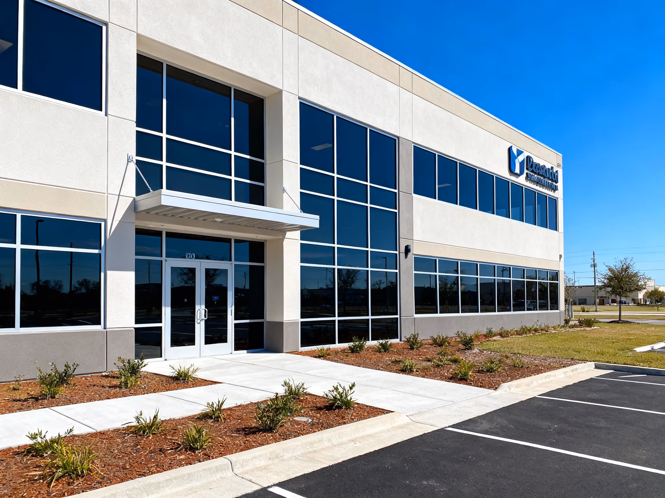 Modern two-story commercial building with large glass windows, a sidewalk, and landscaped plants under a clear blue sky.
