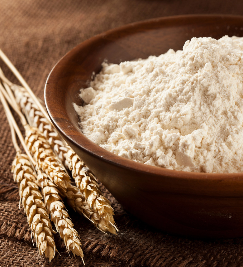 Braune Schüssel mit weißem Mehl neben vier Ähren auf grobem Stoff.