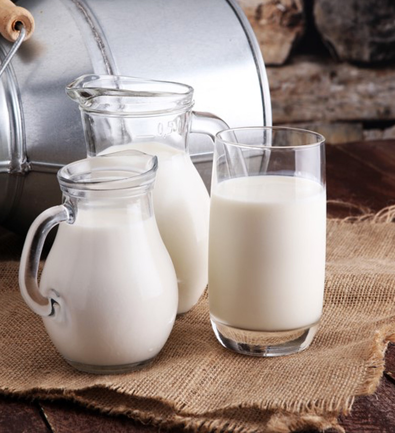 Ein Glas und zwei Krüge mit Milch auf einem rustikalen Stoff auf einem Holztisch, mit einem Metallmilcheimer im Hintergrund.