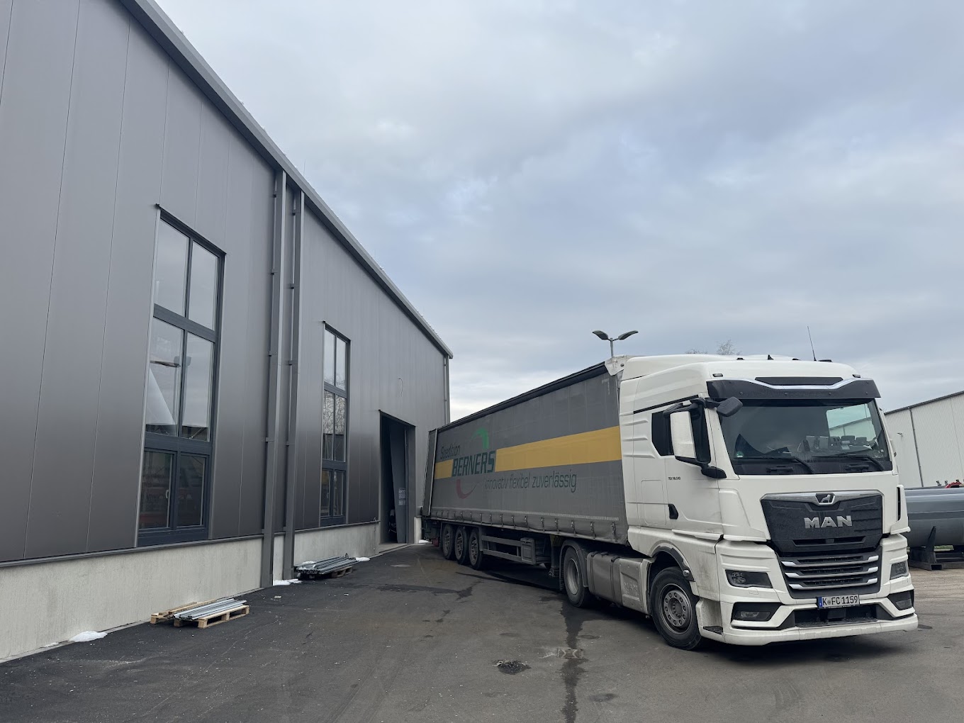 Weißer MAN-Sattelzug mit geschlossener Plane, teilweise vor einem grauen Industriegebäude mit mehreren Fenstern geparkt.